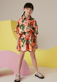 Set de camasa si pantaloni scurti cu model floral in portocaliu, verde si crem; potrivire lejera cu nasturi si talie ajustabila; asortat cu sandale albe cu bretele.