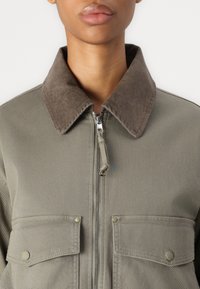 Giacca verde oliva con zip e colletto in velluto a coste marrone, due tasche frontali con pattina e un tessuto textured. Chiusura con zip liscia.