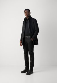 Zwarte trenchcoat met knoopsluiting, gedragen over een zwarte coltrui. Combineert met zwarte fitted broek en enkellaarsjes. Eenvoudig en strak ontwerp.