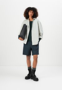 Kvinne med krøllet hår iført en stripete skjorte over en mørk topp, marineblå shorts, grå sokker, svarte militærstøvler, og holder en stor svart veske.