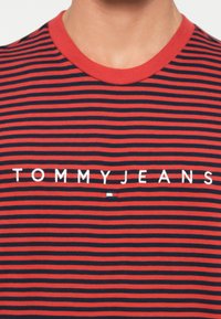 Rød og marineblåstripet T-skjorte med rund hals. Har hvit brodert tekst "TOMMYJEANS" over brystet, med en liten logodetalj.