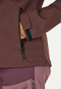 Person’s hand in a brown jacket pocket with a visible zipper, wearing purple pants with zippered pockets, photographed from the side.