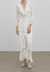 Robe blanche à manches longues avec un col froncé et un motif floral. Présente des détails froncés et une silhouette fluide. Accompagnée de talons à lanières.