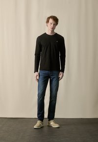 Sort langærmet skjorte, blå denim jeans, lysebrune sneakers. Simpelt design med et lille logo på skjorten. Ensartet baggrund.