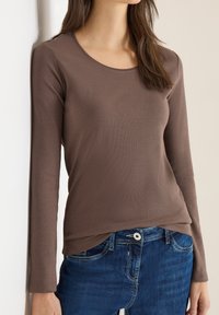 Hnědá košile s dlouhým rukávem z žebrované látky, s kulatým výstřihem a mírně zakřiveným lemem, kombinovaná s modrými džínami.