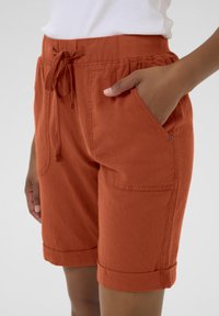 Shorts taille haute, couleur orange rouille, fabriqués en tissu texturé, dotés d'une taille élastique avec cordon de serrage et de poches latérales avec des ourlets à revers.