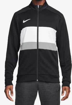Man draagt een zwart jack met ritssluiting, witte en grijze horizontale strepen en een wit Nike-logo op de borst.