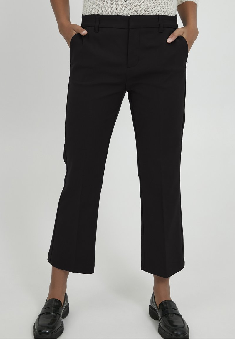 Personne portant un pantalon noir droit coupé court, les mains dans les poches et des mocassins noirs, debout sur un fond clair uni.