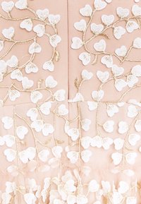 Vyšívaná tkanina má květinové vzory s bílými okvětními lístky a zlatou nití na jemně růžovém pozadí, což ukazuje jemný, strukturovaný vzor.