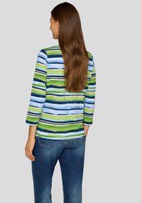 Frau mit langen braunen Haaren, die ein gestreiftes blau-grün-weißes Hemd und blaue Jeans trägt, von hinten vor einem grauen Hintergrund gesehen.