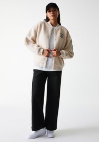 Beige fleece bomber jacket with ribbed cuffs, featuring a logo on the chest. Paired with black wide-leg pants and white sneakers. Black cap.