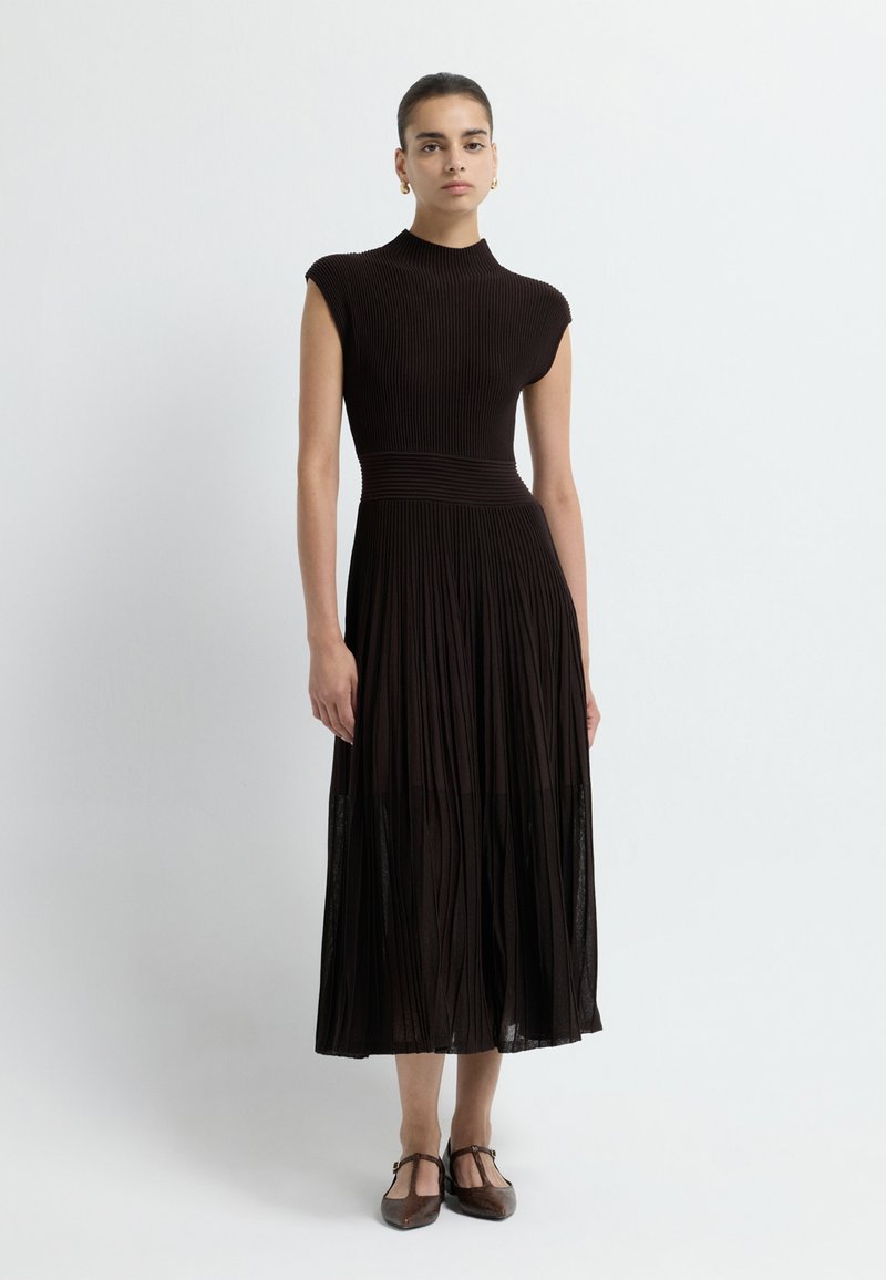 Texturované, žebrované černé šaty s vysokým výstřihem, upnutou korzetovou částí a skládáním v rozšířené sukni. Lem obsahuje průsvitné detaily.