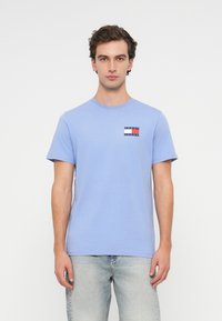 T-shirt de algodão azul claro com gargantinha redonda. Apresenta um pequeno logótipo retangular em vermelho, branco e azul marinho no peito. Mangas curtas.