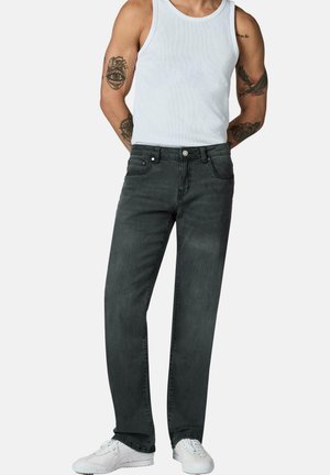 Mann trägt schwarze Straight-Leg-Jeans, weißes Tanktop und weiße Sneakers, mit sichtbaren Arm-Tattoos, vor neutralem Hintergrund stehend.