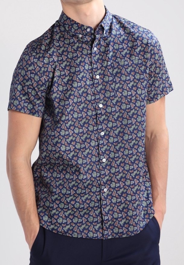 Homme portant une chemise bleu marine à manches courtes avec un petit motif paisley multicolore, mains dans les poches d’un pantalon sombre.