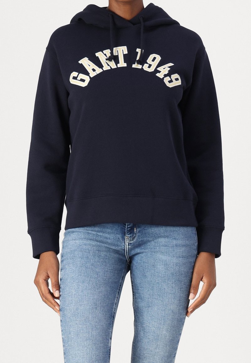 Personne portant un sweat à capuche bleu marine GANT 1949 et un jean bleu clair sur un fond blanc uni.