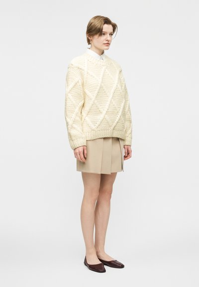 Άτομο που φοράει εκρού πουλόβερ με πλεκτό σχέδιο, μπεζ πλισέ φούστα, λευκό πουκάμισο και σκούρα καφέ φλατ παπούτσια, στέκεται μπροστά σε λευκό φόντο.