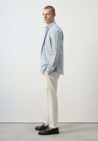 Homme debout de profil portant une chemise à carreaux bleu clair, un pantalon blanc et des mocassins noirs, les mains dans les poches sur un fond uni.