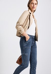 Beige verkürzte Jacke mit einer figurbetonten Silhouette über einem weißen Oberteil, kombiniert mit dunkelblauen Denim-Cargo-Hosen mit Seitentaschen.