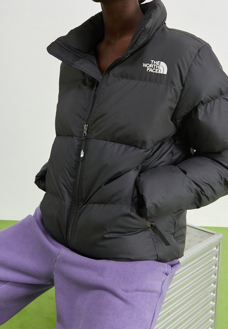 Czarna kurtka puchowa z wysokim kołnierzem, zamkiem błyskawicznym i logo The North Face na lewej piersi. Połączona z fioletowymi spodniami dresowymi.