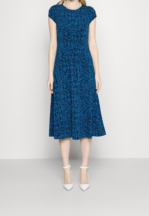 Femme portant une robe bleue et noire à motifs, longueur genou, avec des manches courtes et des talons blancs à bride à la cheville, debout devant un fond clair uni.