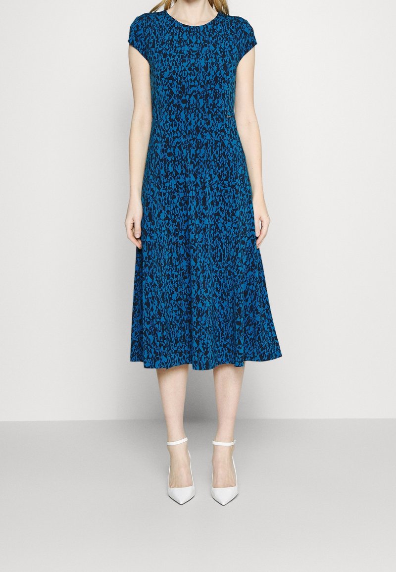 Femme portant une robe bleue et noire à motifs, longueur genou, avec des manches courtes et des talons blancs à bride à la cheville, debout devant un fond clair uni.