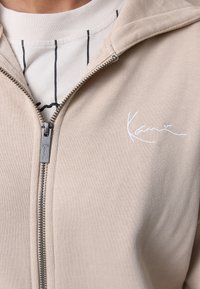 Light beige zip-up jacket with a textured finish, featuring a white embroidered logo. Layered over a white shirt with vertical black stripes.