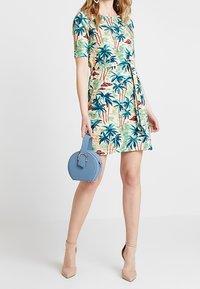Šaty s tropickým palmovým potiskem v zelené, modré a oranžové barvě; krátké rukávy a rozšířená sukně. Ladící kulatá modrá kabelka. Béžové vysoké podpatky.