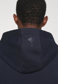 Felpa navy con cappuccio strutturato e logo discreto sul retro. Realizzata in un tessuto morbido e texturizzato, che sottolinea la semplicità nel design.