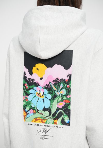 Siva kapuca s velikim, barvnim grafičnim motivom, ki vključuje cvet, metulja in sonce, ter besedilo "MARC JACOBS ARTIST CAPSULE."