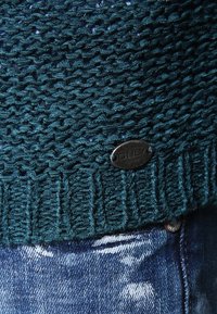 Sweter w kolorze teal z dzianiny, z teksturowanym wzorem i ściągaczem na końcu, ozdobiony metalową metką z logo. Nosi się go na wyblakłe niebieskie dżinsy.