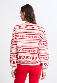 Czerwony i biały puszysty sweter z dzianinowymi wzorami serc, kokardek i płatków śniegu, z wyraźnie wyeksponowanym napisem "RADOSNY".