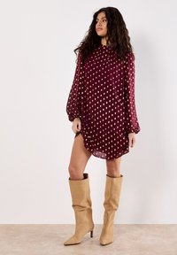 Donna con lunghi capelli ricci che indossa un vestito bordeaux con pois dorati e stivali alti fino al ginocchio in camoscio beige, in piedi contro un muro semplice.