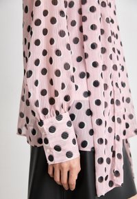 Blouse sheer rose clair avec de grands pois noirs, poignets boutonnés, portée avec une jupe noire, gros plan sur le bras et la main.