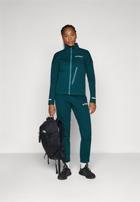 Veste et pantalon de sport teal avec poches zippées et détails réfléchissants. Sac à dos noir avec logo sur l'épaule. Baskets noires.
