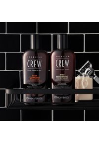 Kaksi tummaa amberpullollista American Crew Daily Cleansing Shampoo -shampoota ja Daily Moisturizing Conditioner -hoitoainetta mustilla etiketeillä mustalla hyllyllä.