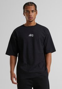 Sort kortærmet t-shirt lavet af bomuld. Har et lille hvidt grafisk design på midten af brystet. Løs pasform med bådudskæring.