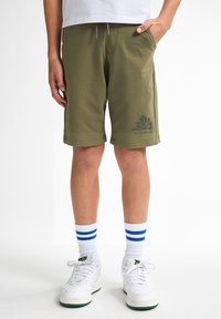 Shorts vert olive avec un design de logo, taille à cordon de serrage, et poches latérales, associés à des chaussures blanches et des chaussettes rayées bleues.