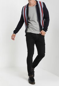 Gilet marine avec des rayures multicolores verticales sur un t-shirt gris, associé à un jean noir et des mocassins noirs, offrant un look décontracté.