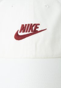 Biely baseballový klobúk vyrobený z bavlny so štruktúrovaným tvarom. Obsahuje bordový vyšívaný logo "Nike" a viditeľné detaily šitia.