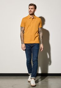 Żółta koszulka polo z krótkim rękawem i kołnierzykiem, w połączeniu z ciemnoniebieskimi dżinsami i białymi trampkami. Zawiera skomplikowane tatuaże na obu ramionach.