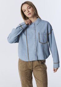 Lichtblauwe denim shirt met een boxy fit, contrasterende bruine stiksels, een borstzak en knoopsluiting aan de voorkant. Gepaard met tan suede broek.