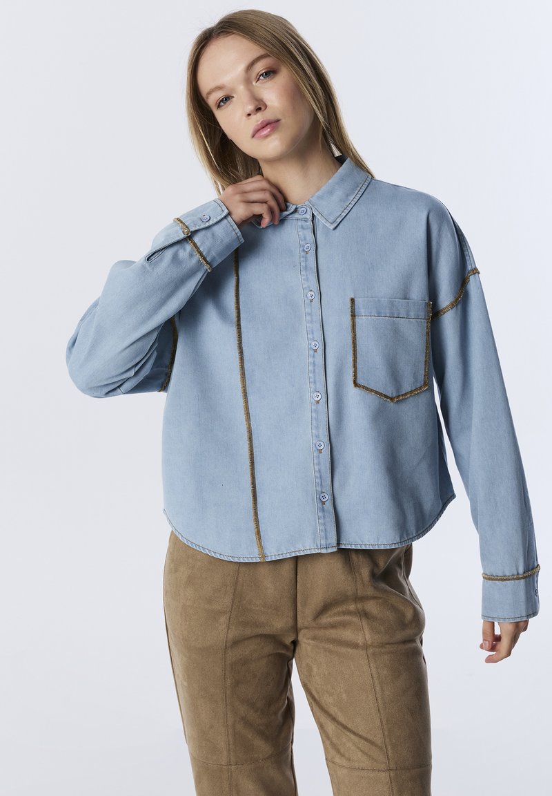 Lichtblauwe denim shirt met een boxy fit, contrasterende bruine stiksels, een borstzak en knoopsluiting aan de voorkant. Gepaard met tan suede broek.