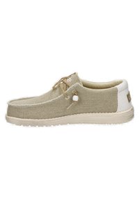 Zapatilla de lona beige con parte superior texturizada, puntera redonda, suela de goma beige y cordones decorativos que presentan una pequeña perla blanca.