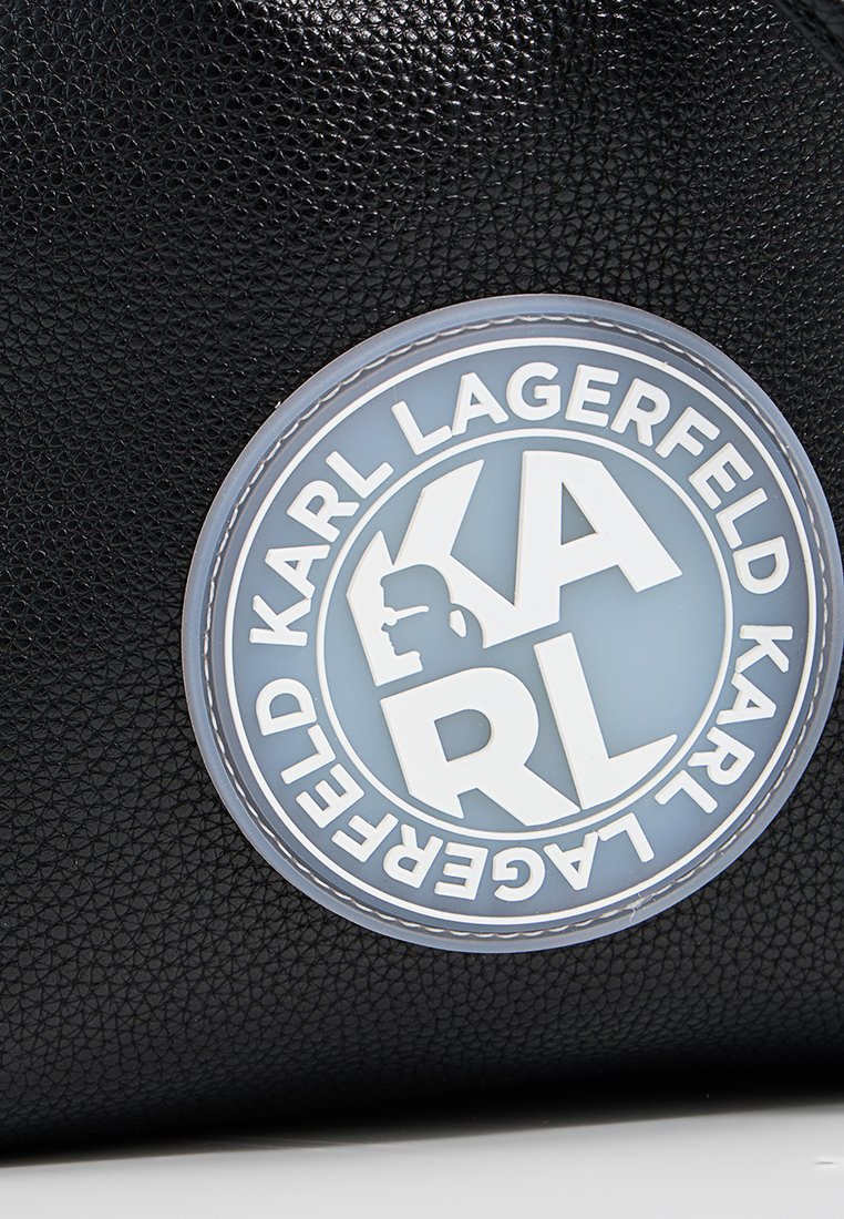 Schwarz strukturiertes Lederhandtasche mit einem runden hellblauen Logo-Patch und weißer Schrift, einschließlich "KARL LAGERFELD" und einem Profil-Silhouette.