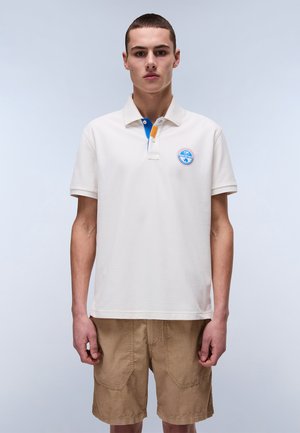 Creme polo skjorte med blå og orange krave detalje, med et rundt blåt logo på brystet. Modellen bærer brune shorts.