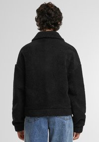 Zwarte fleece jas met een gestructureerde afwerking, een brede kraag en een relaxte pasvorm. Gecombineerd met lichtblauwe hoge taille jeans, gezien van achteren.