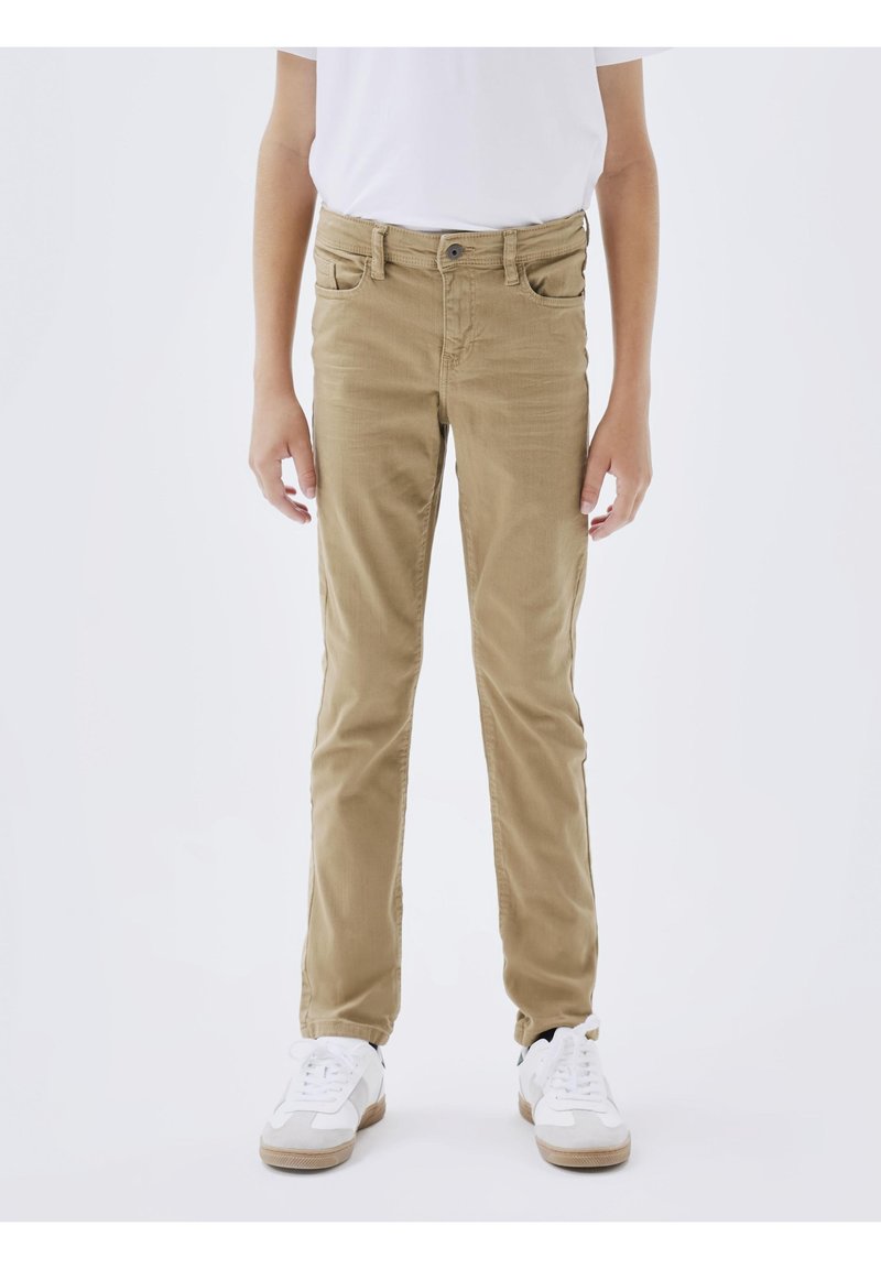 Enfant portant un pantalon beige slim et des baskets blanches, debout sur un fond blanc uni.