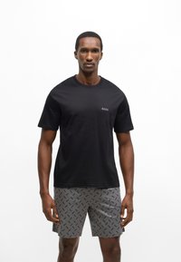 Μαύρο κοντομάνικο μπλουζάκι με διακριτικό λογότυπο "BOSS" στο στήθος, συνδυασμένο με γκρι σορτς που έχουν επαναλαμβανόμενο μοτίβο με το όνομα της μάρκας.