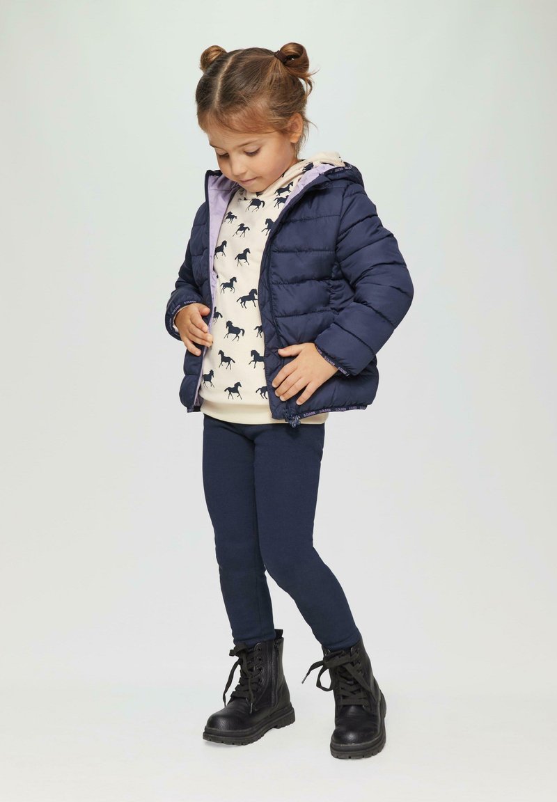 Marineblaue Steppjacke mit Kapuze, beiger Pullover mit schwarzem Pferdemuster, dunkle Leggings und schwarze Schnürstiefel. Glatte Textur, lässiges Design.